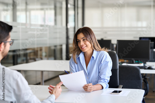 Middle aged Hispanic business woman financial advisor, bank manager consulting client working in office. Professional female hr executive having job interview meeting with male recruit employee. photo