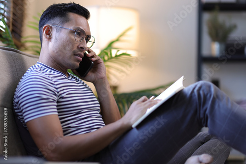 Man reading a book and writing a diary while relaxing on the sofa at home.