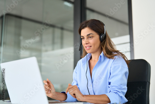 Busy mature business woman call centre representative customer support agent talking to client, smiling middle aged senior female operator wearing headset working using laptop computer in office.