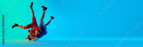 Two young male athletes in motion, wrestling, fighting against blue background in neon light. Concept of combat sport, martial arts, competition, tournament, athleticism. Banner for ad, space for text photo