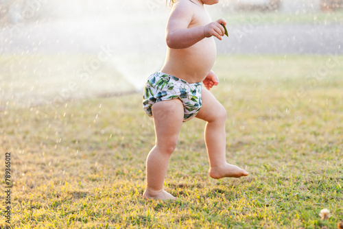 toddler in cloth nappy running under sprinkler in yard on hot afternoon photo