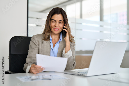 Middle aged Hispanic professional business woman executive making call having conversation at work. Mature female manager or entrepreneur talking on the phone checking document sitting at office desk.