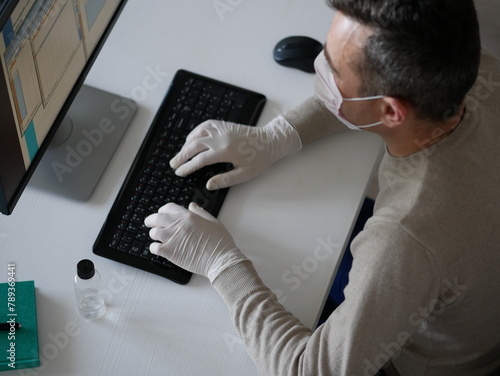 Young woman wearing protective gloves on hands and mask on face working from office or home using laptop on desk.