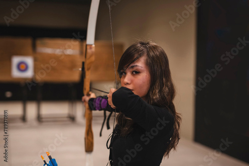 A young Asian woman glances at the camera while at an indoor archery range, bow in hand. She wears an arm guard, embodying focus and precision while indulging in her hobby. photo