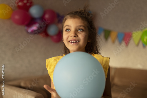 Smiling real girl 7 years old in yellow dress with blue balloon against festive backdrop in holiday at home