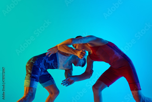 Two men, wrestling athletes grapple in wrestling match, showing strength and endurance against blue background in neon. Concept of combat sport, martial arts, competition, tournament, athleticism photo