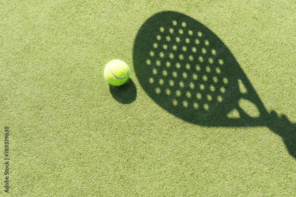 Obraz premium Shadow of a padel racket with a yellow ball on the green grass.
