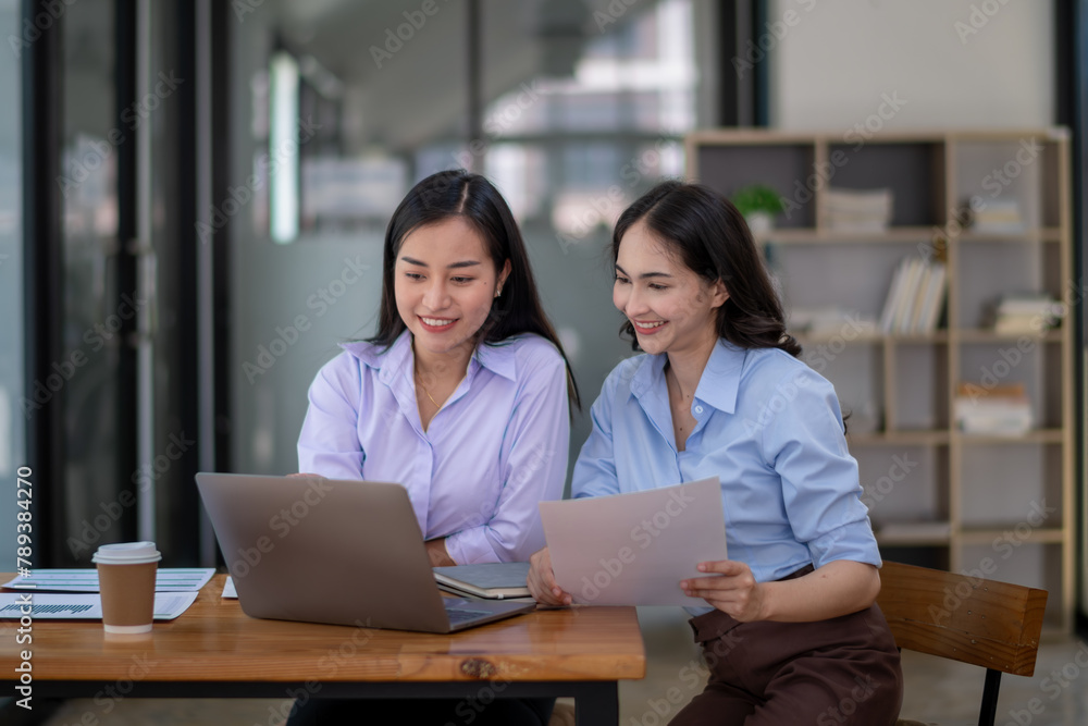 Two smiling businesswomen collaborating in a modern office environment, reviewing a document and using a laptop. Two smiling businesswomen collaborating in a modern office environment, reviewing a document and using a laptop.