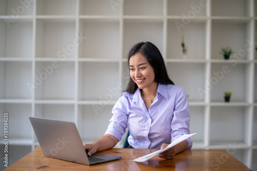 Smiling Asian businesswoman working at her desk with laptop, surrounded by minimalist style shelving in a contemporary office.