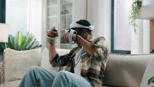 African American man player enjoying at home couch playing vr game fight boxing ethnic guy gamer male play online fighting wear headphones virtual reality 3d helmet glasses hold controllers joysticks photo