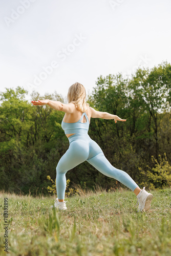 Side view of an athletic girl performing forward lunges in the park. Beautiful blonde Caucasian woman in blue tight tracksuit. Blonde girl at an outdoor training session