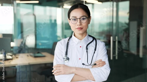 Portrait of young serous asian female doctor in white coat and glasses looking at camera in modern hospital clinic. Confident woman medical worker physician poses with crossed arms standing in office photo
