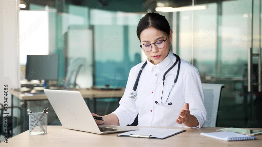 Young asian female doctor in white coat talking on video call using laptop computer in modern hospital clinic. Medical worker physician consults a patient sitting at a desk at a workplace in an office Young asian female doctor in white coat talking on video call using laptop computer in modern hospital clinic. Medical worker physician consults a patient sitting at a desk at a workplace in an office