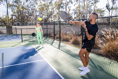 Co-ed pickleball players on a court outside in the summer. Both athletes are fit and the man and woman holding their paddle are each ready to play. 