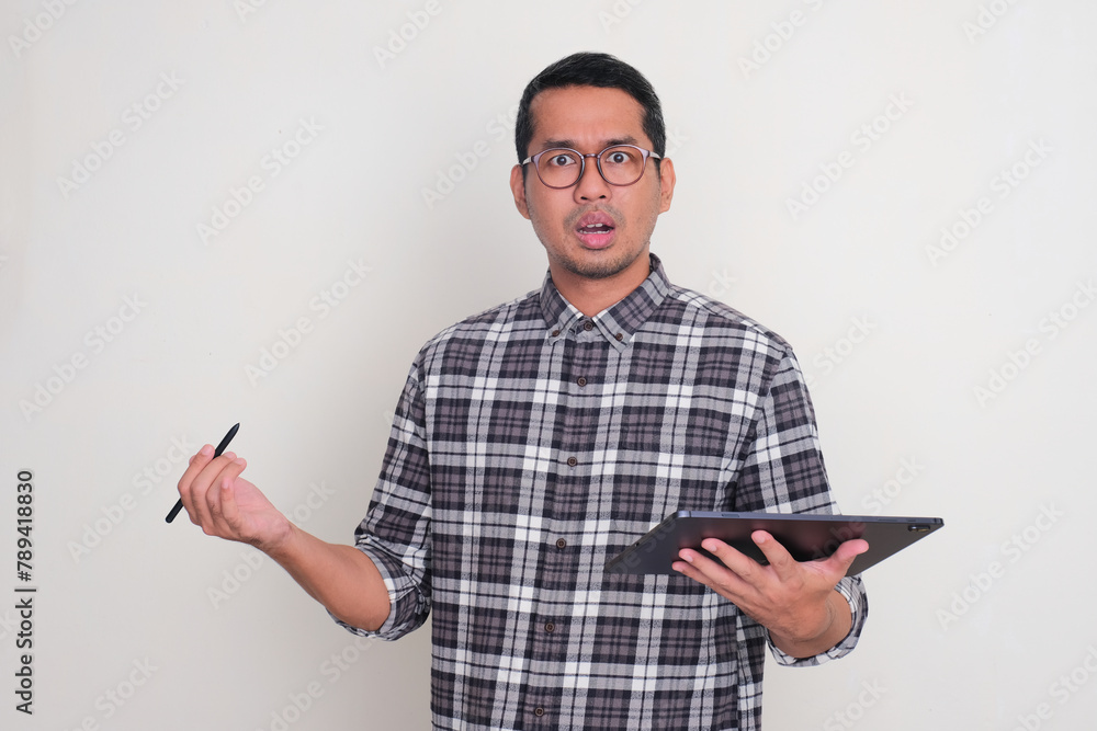 Adult Asian man holding mobile tablet device with confused face expression Adult Asian man holding mobile tablet device with confused face expression