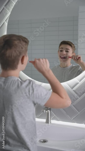 Happy boy cleaning teeth in white bathroom looking to mirror. Healthy child makes everyday morning dental care routine. Brushing teeth with toothbrush, caring about dental hygiene at home. Vertical