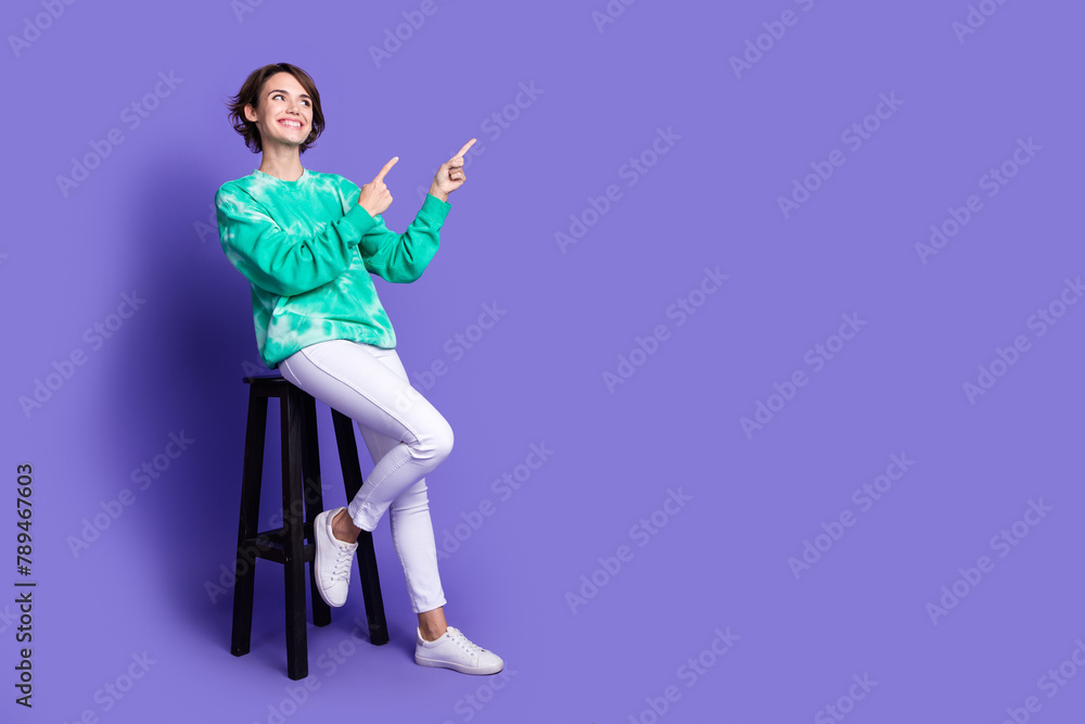 Full size photo of attractive woman sit chair point look empty space dressed stylish green clothes isolated on violet color background Full size photo of attractive woman sit chair point look empty space dressed stylish green clothes isolated on violet color background