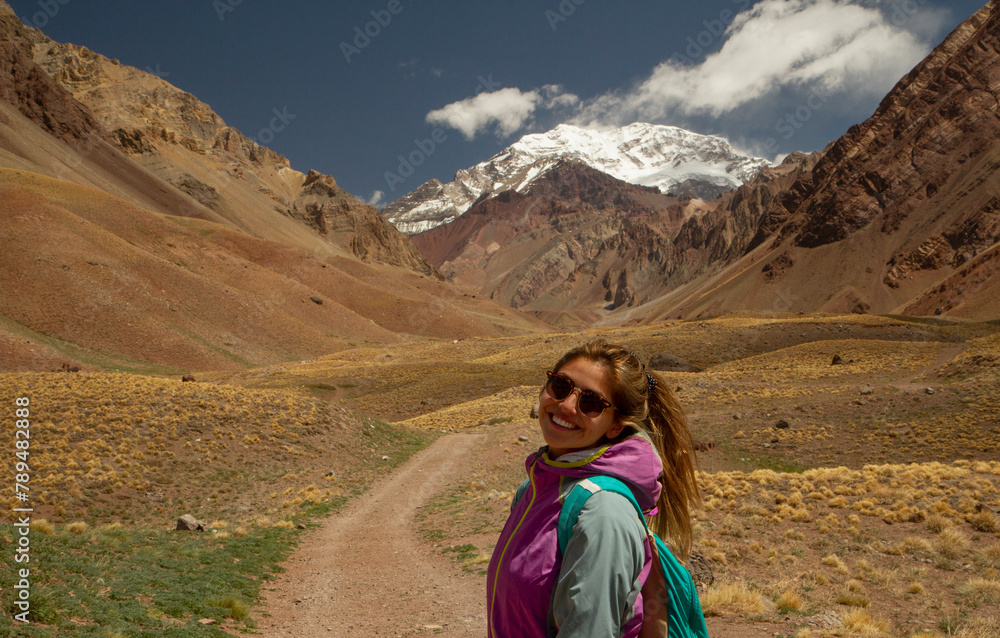 View of a young woman wearing sunglasses, hiking along the dirt road leading to Aconcagua mountain in Mendoza.