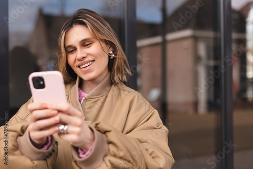 Attractive blonde woman wear bomber hold mobile phone and laughing, look happy and smiling. Amazing enjoy woman walk near glass office. Girl read fun sms, news or joke.