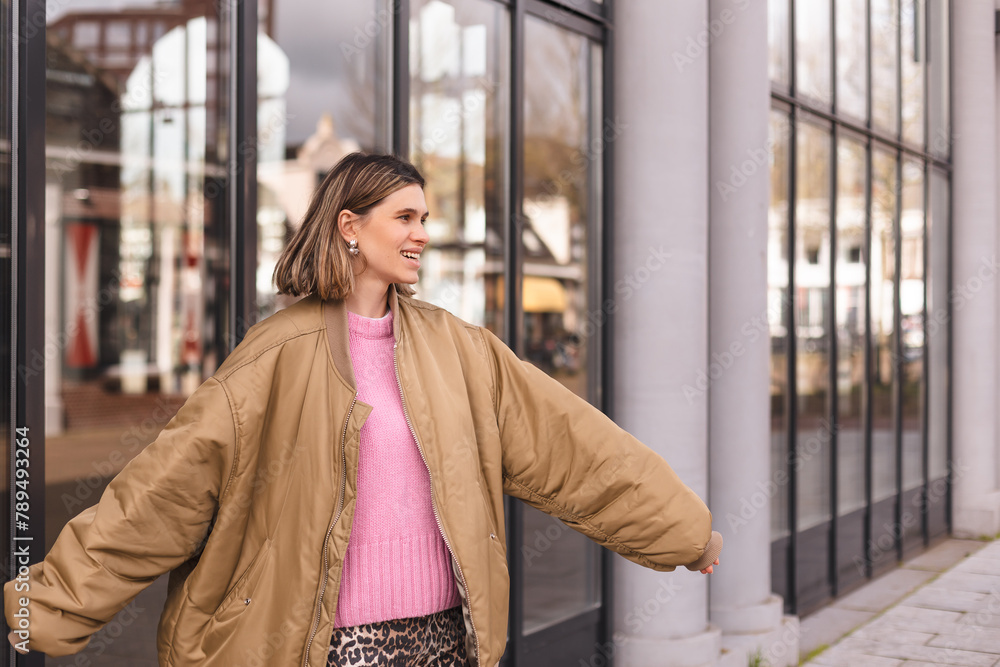 Fototapeta premium Happy blonde short hair woman wear bomber jacket walking in the street and smiling to camera. Stylish girl in fashion outfit walking over the city. What a beautiful day out. Girl turn around.