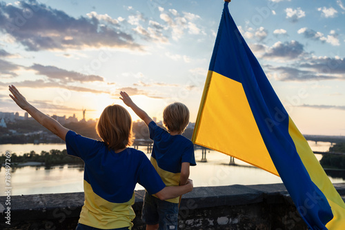 Mom and son with the Ukrainian flag stand high on the roof against the background of the sky and the Dnieper River in Kyiv. Patriotism, drawing attention to the war in Ukraine. Independence Day. photo