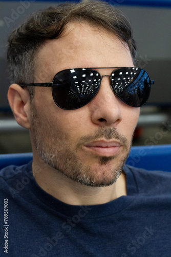 Closeup view of a white caucasian man in his 30s wearing sunglasses that reflect the room lights. 