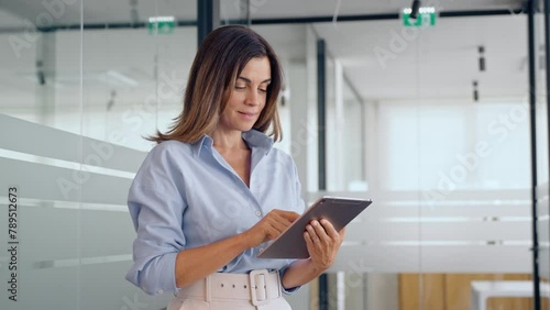 Smiling mature professional business woman executive using digital tablet at work. Busy middle aged businesswoman manager leader entrepreneur working on tab tech device standing in corporate office.