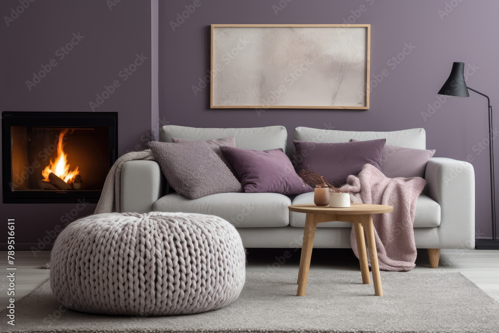 Fototapeta premium Knitted pouf near grey sofas with purple pillows. Fireplace against grey wall with poster frame. Scandinavian home interior design of modern living room