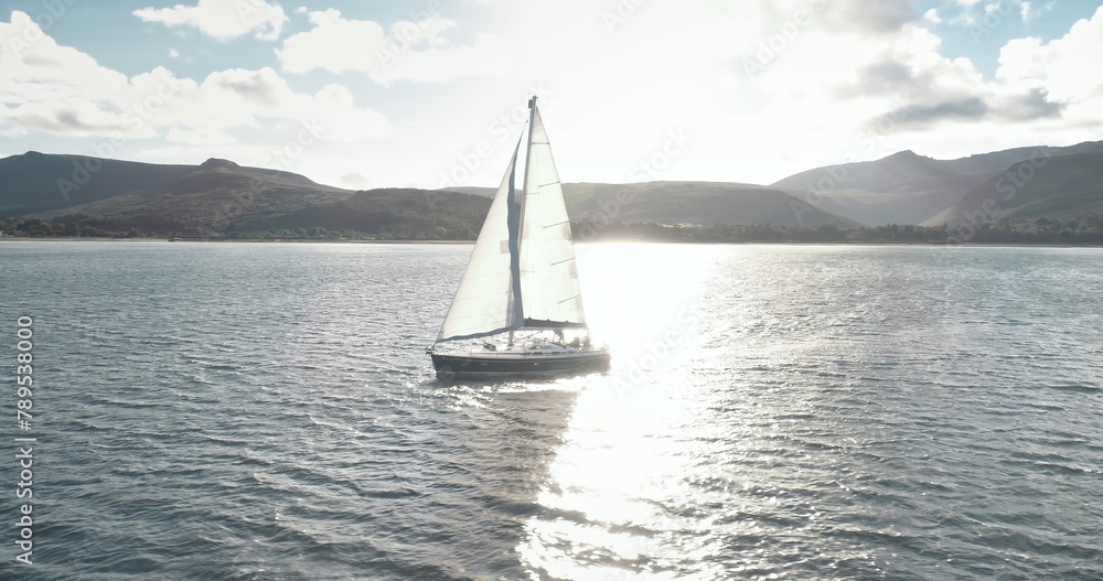 Slow motion yacht racing at ocean bay with highlang island coast aerial. Sun reflect at sailboat. Sunlight reflection at sea shore of Arran isle, Scotland, Europe. Luxury ship at summer cruise