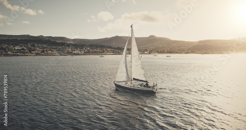 Sun seascape with white sailboat aerial. Mesmerezing landscape with sail boat at summer. Sunlight over sail boat at ocean bay. Sea shore of Arran Island with vessel. Serene water scenery drone shot