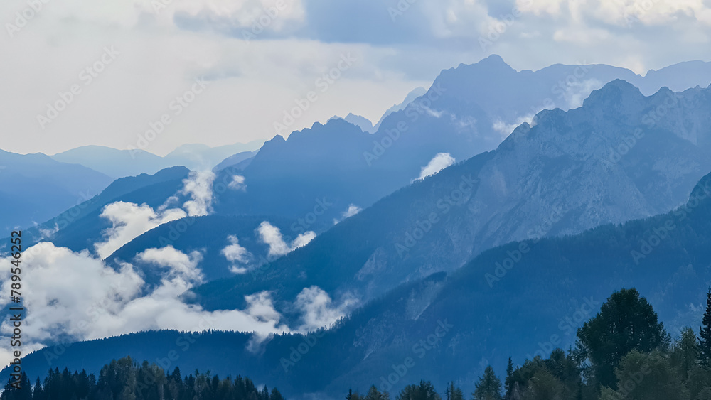 Panoramic view from Monte Lussari in Camporosso, Friuli Venezia Giulia, Italy. Looking at majestic mountain peaks of Julian Alps and Karawanks, border to Austria Slovenia. Misty atmosphere in nature