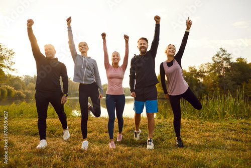 Group of happy fit sporty slim confident motivated diverse multiethnic athletes in sportswear standing on green river bank in park, raising hands up and smiling. Nature, sport, outdoor workout concept