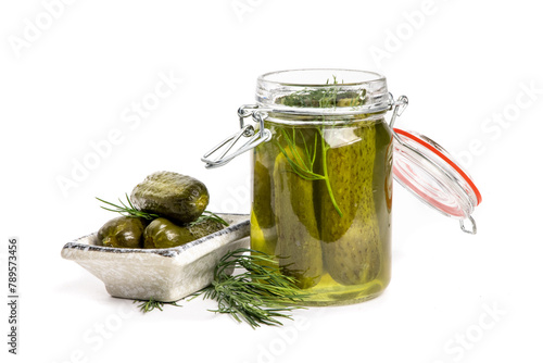 A glass home canning jar of baby dill pickles with the lid open and pickles on a small plate showing the dill herb isolated on white