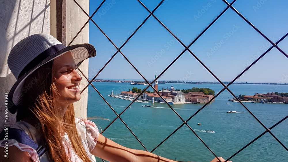 Tourist woman wearing hat with aerial view from St Mark bell tower Campanile of old town Venice, Veneto, Italy, Europe. Looking at San Giorgio Maggiore island, Venetian lagoon. UNESCO World Heritage - obrazy, fototapety, plakaty 