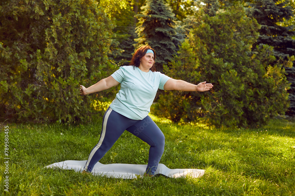 Full length body shot of confident motivated fat stout overweight woman in sportswear doing yoga exercises in nature and standing in Virabhadrasana B Warrior pose on green grass in lush summer park Full length body shot of confident motivated fat stout overweight woman in sportswear doing yoga exercises in nature and standing in Virabhadrasana B Warrior pose on green grass in lush summer park