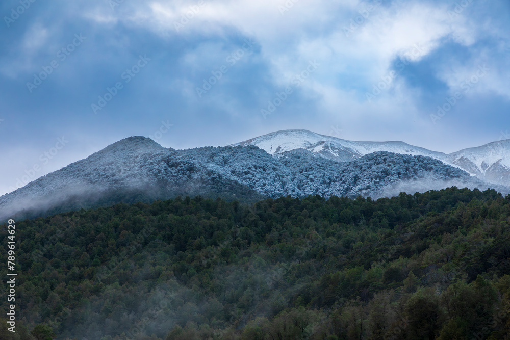 Fototapeta premium Photograph of cloud covered snow capped mountains with lush foliage in Fiordland National Park on the South Island of New Zealand