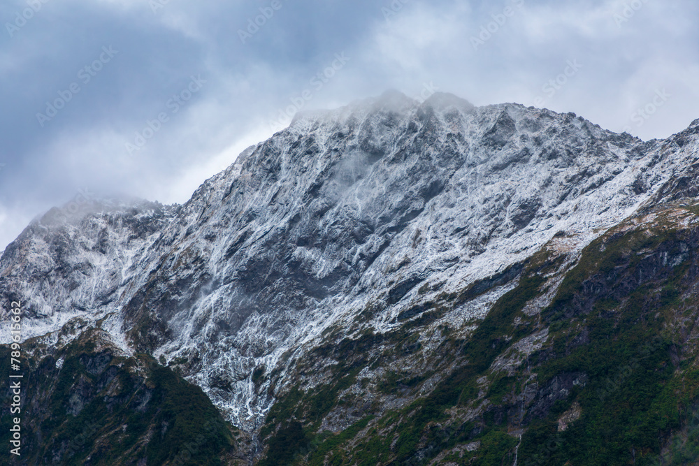 Photograph of cloud covered snow capped mountains with lush foliage in Fiordland National Park on the South Island of New Zealand