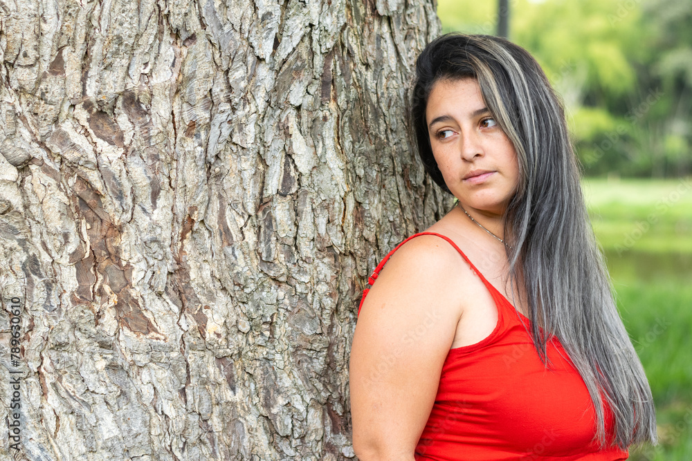 latina girl leaning against a big tree in a red dress, looking sadly to one side latina girl leaning against a big tree in a red dress, looking sadly to one side