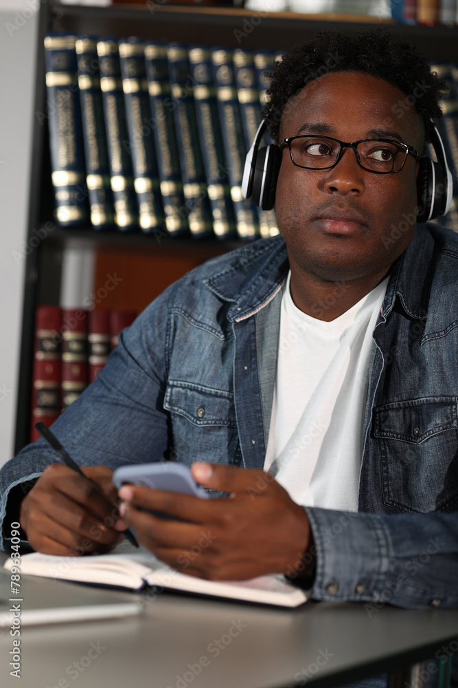 Close-up of a young african employee at his desk in a modern office space. Cute thoughtful serious african man in denim shirt sitting in headphones holding mobile phone in hand