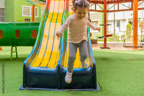 Schilderij op canvas Una linda niña de dos años sonriente juega en un tobogan dentro del patio de un condominio