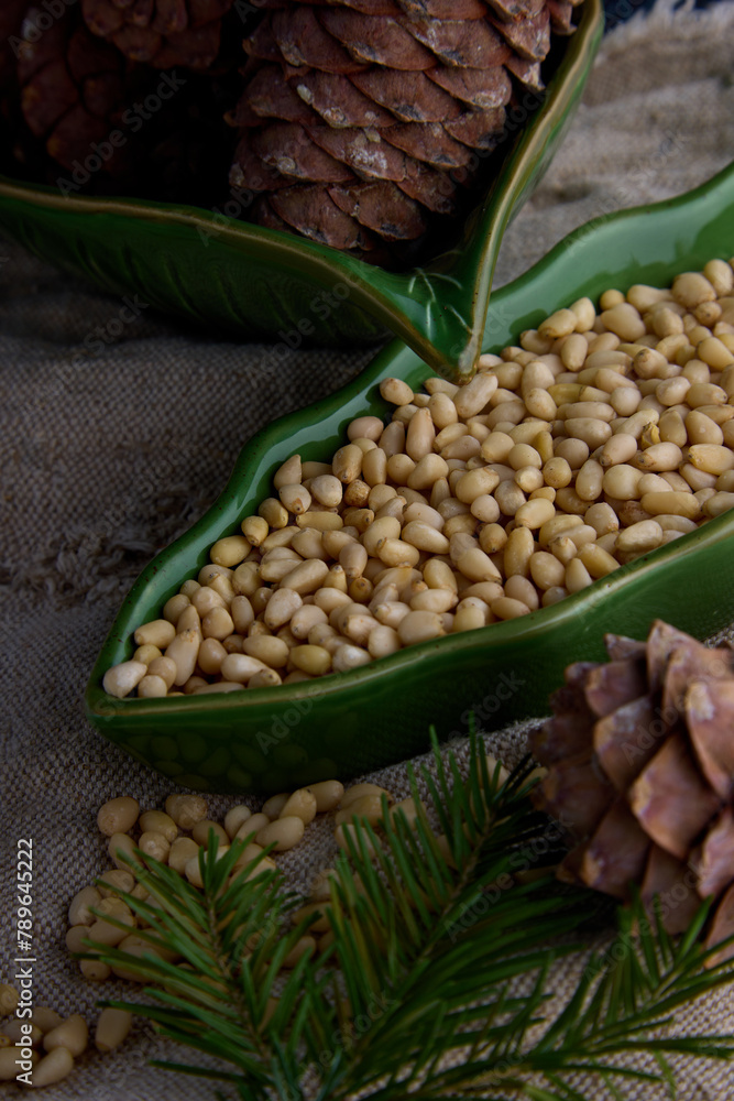 Lot of peeled pine nuts in a green plate in the shape of a leaf on burlap. Cedar cones and a coniferous branch on the background of old burlap in a ceramic plate Lot of peeled pine nuts in a green plate in the shape of a leaf on burlap. Cedar cones and a coniferous branch on the background of old burlap in a ceramic plate