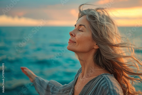 Free happy middle aged woman with arms outstretched and eyes closed enjoying fresh air on the beach at sunset, meditation and unity with nature