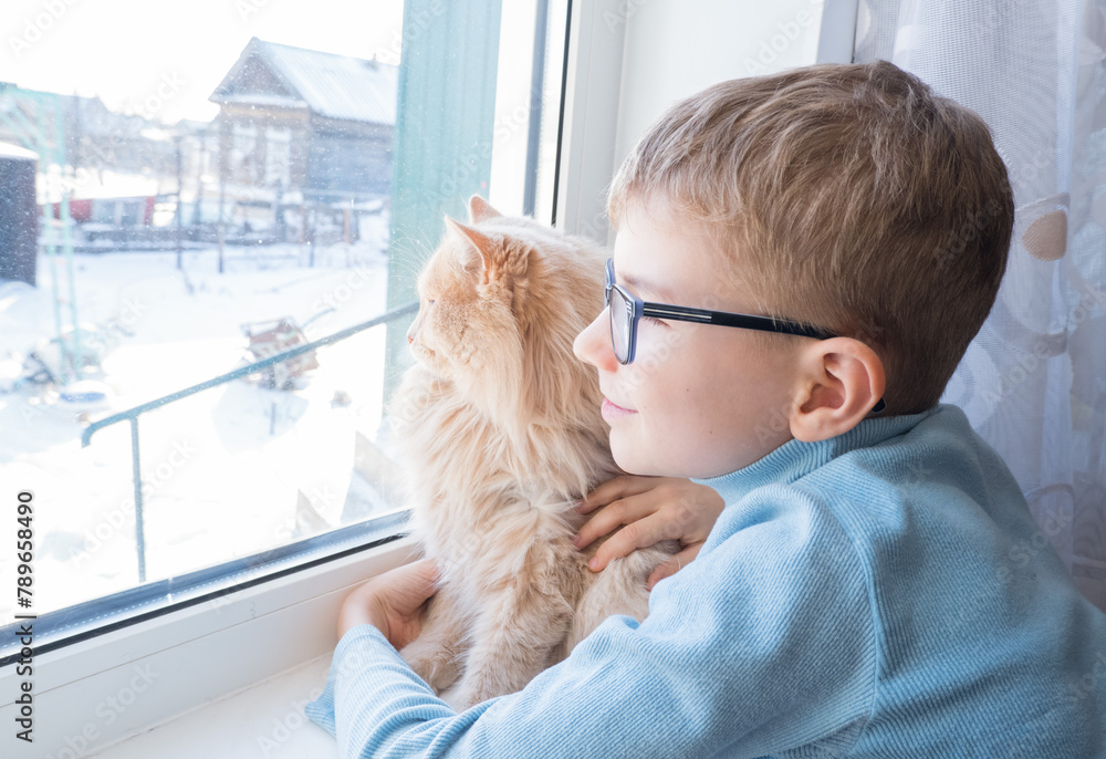 Beautiful scene two friends looking out fo window. Small boy with a cat stares out of window watching falling snow, focus on cat