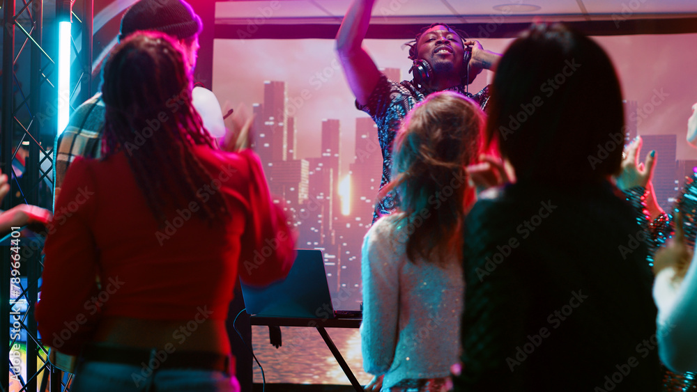 Young man clubbing with people at bar, having fun with DJ mixing station on nightclub stage with disco ball. Funky person creating live performance on audio panel, spotlights at club.