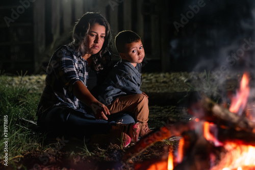 young latin woman carrying her son, sitting in front of a campfire at night, looking sad and bored.