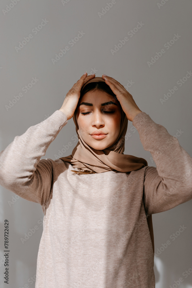 Smiling young female in hijab touching head while standing in studio Smiling young female in hijab touching head while standing in studio