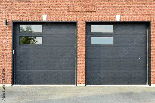 Exterior of a red brick residential building with double black metal car garage doors. There are two small glass panels in each solid door. The driveway is concrete. There's a lit fixture on the wall.