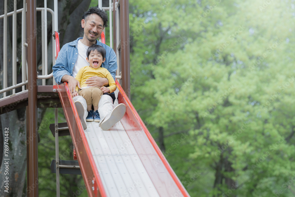 Obraz premium 新緑の公園の滑り台で遊ぶアジア人（日本人）の親子 2人家族 パパ(父親）の子育てや育児の休日のイメージ