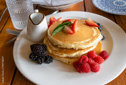 stack of pancakes with strawberries, blackberries, and raspberries on a white plate with a small syrup jug on a table in a restaurant