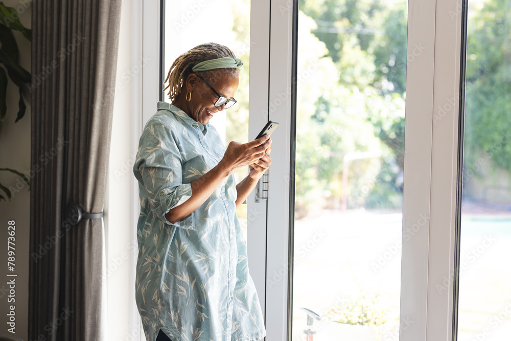 African American senior woman standing by window at home, looking at smartphone, copy space African American senior woman standing by window at home, looking at smartphone, copy space